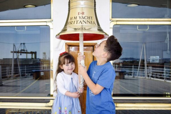 A boy, wearing a blue t-shirt  stands beside a girl wearing a blue dress and white cardigan both hold on to the rope coming from a silver bell with red interior. The bell says HM Yacht Britannia on it.,© Helen Pugh