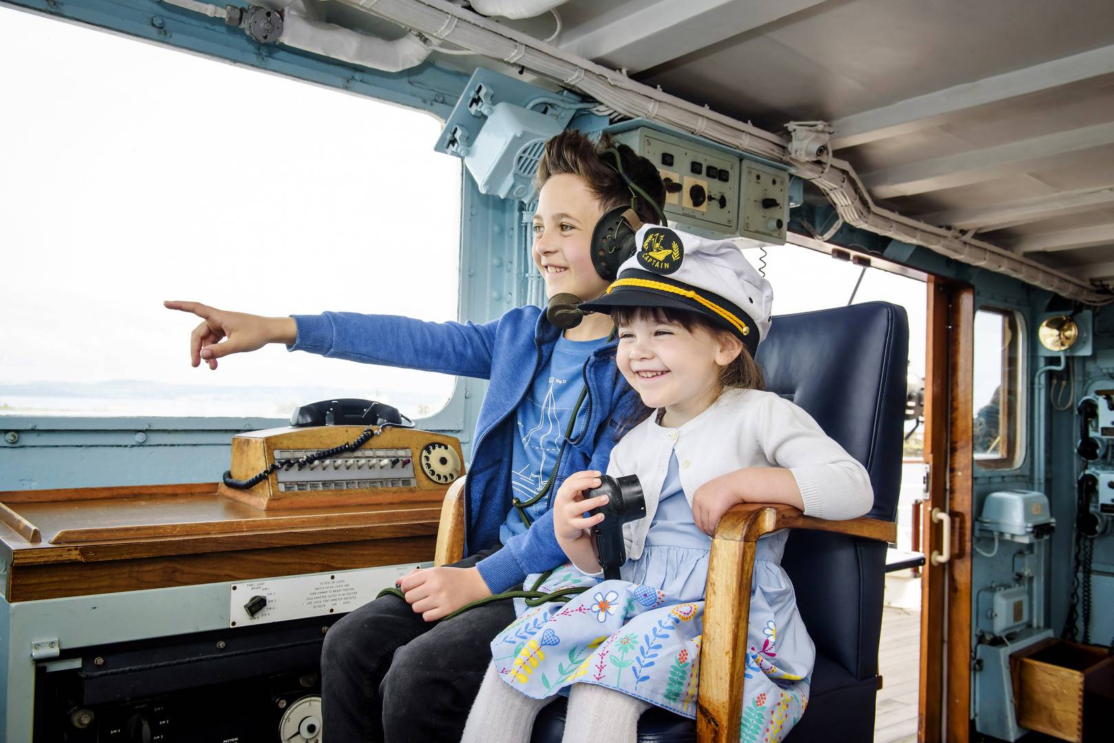 A boy, wearing a blue t-shirt and blue hooded jumper, sits beside a girl wearing a blue, flower patterned dress, white cardigan and captain's hat sit in the captain's chair in the ship's bridge. ,© Helen Pugh