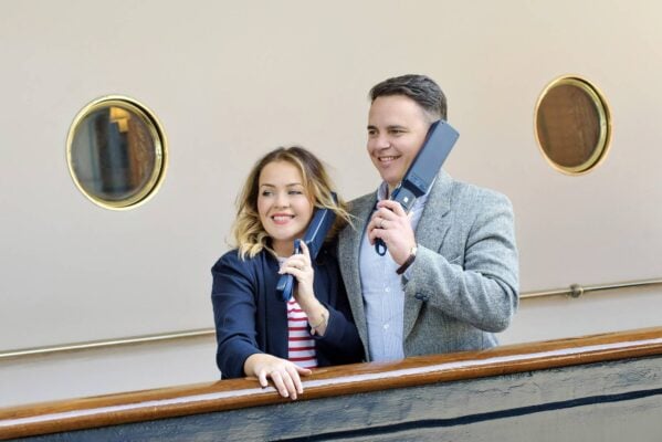 A woman wearing a navy blazer with a white and red striped t-shirt and her husband, who wears a blue and white shirt with grey woollen blazer both listen to audio handsets on the deck., Helen Pugh