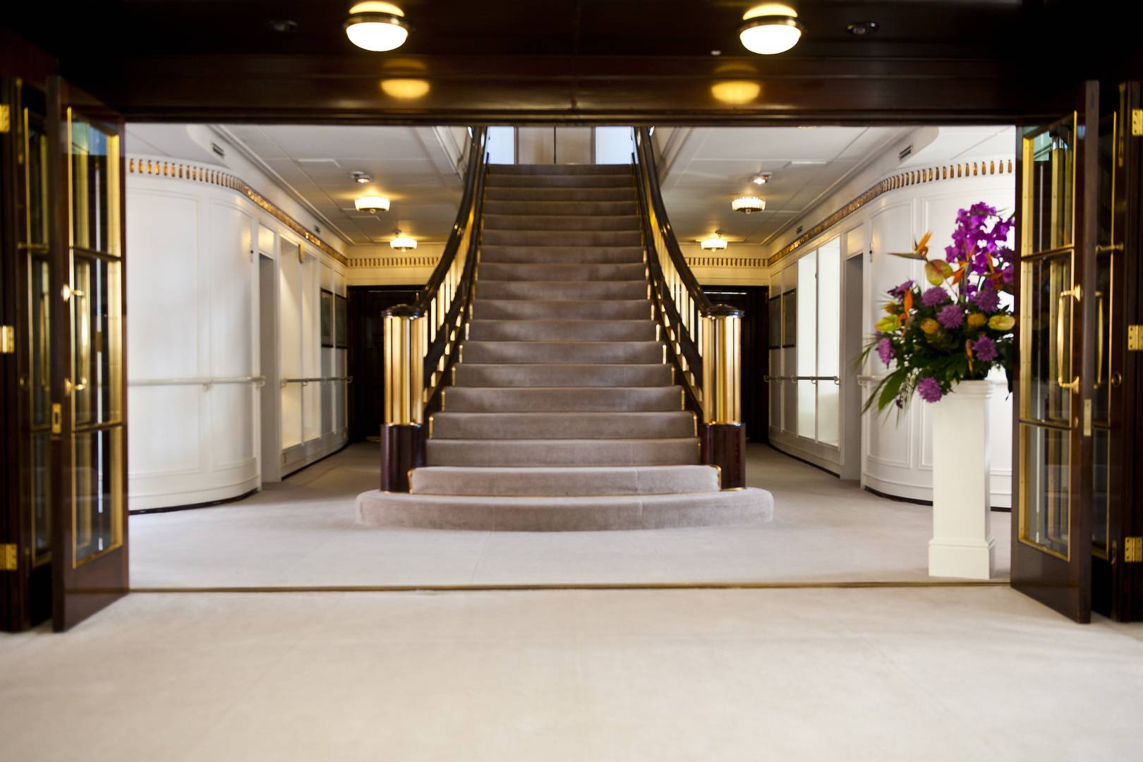 The Grand Staircase, gold and dark wood railings with a taupe coloured carpet. Pink and yellow flowers are on a white podium to the right of the staircase.,© Marc Millar