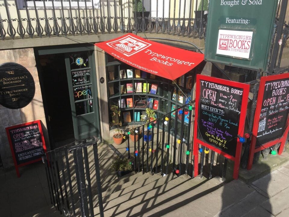 The frontage of Typewronger Books on a sunny day, there are blackboards advertising our opening hours and the next event, colourful drawings on the windows, and a large red sun shade over the window emblazoned with our logo and website details.,© Tee Hodges