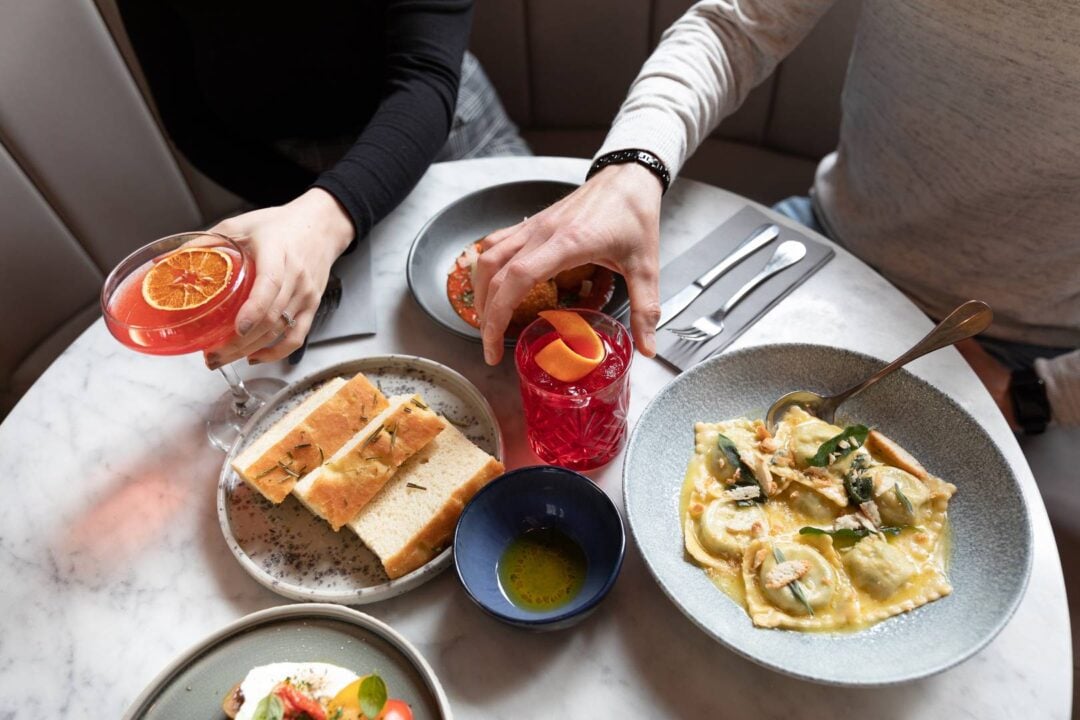 Image of marble table covered in cocktails, ravioli, side focaccia, arancini