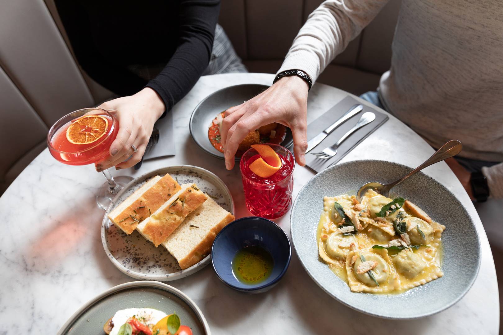 Image of marble table covered in cocktails, ravioli, side focaccia, arancini