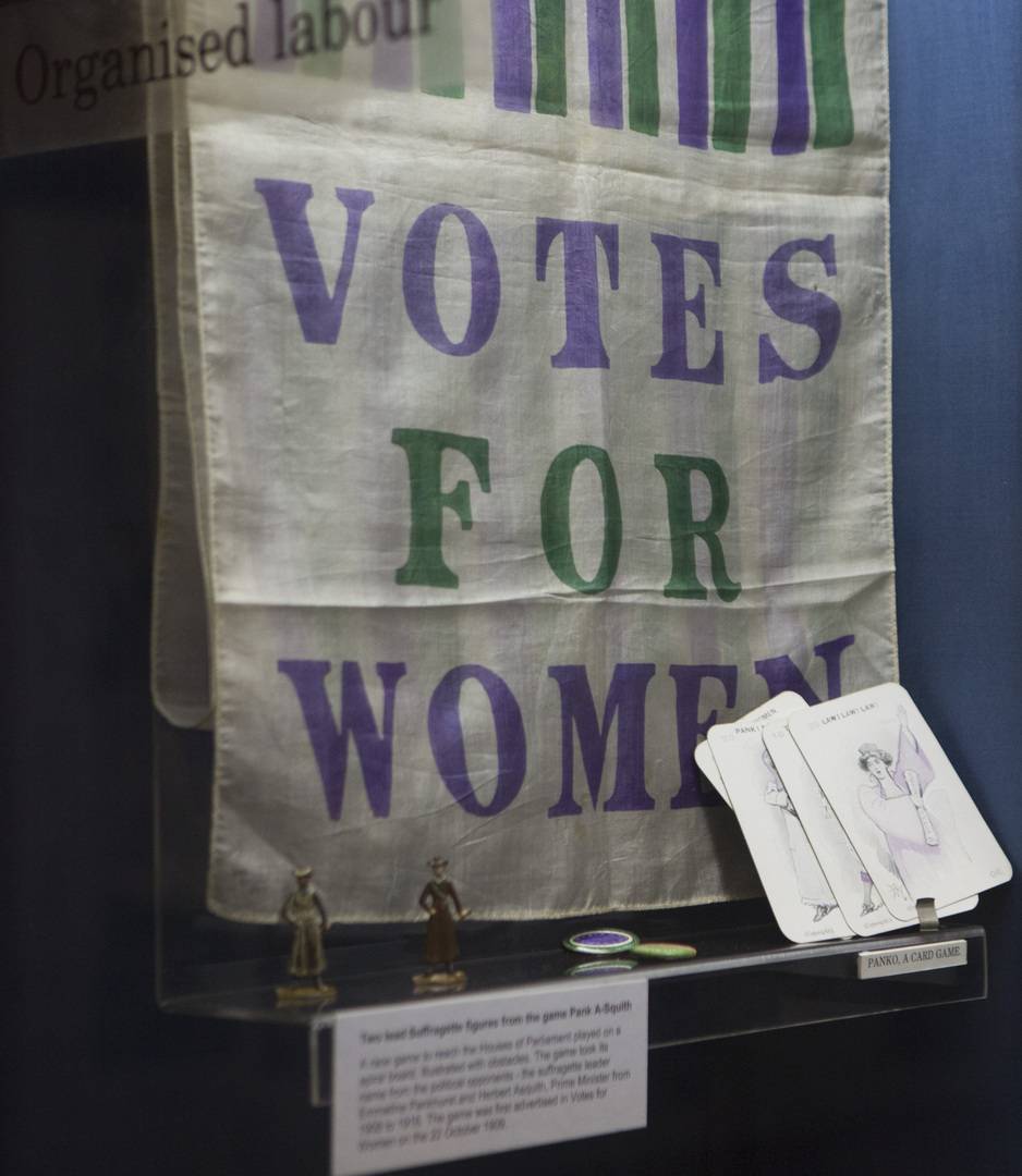 'Votes For Women' flag in purple and green font displayed in glass case.