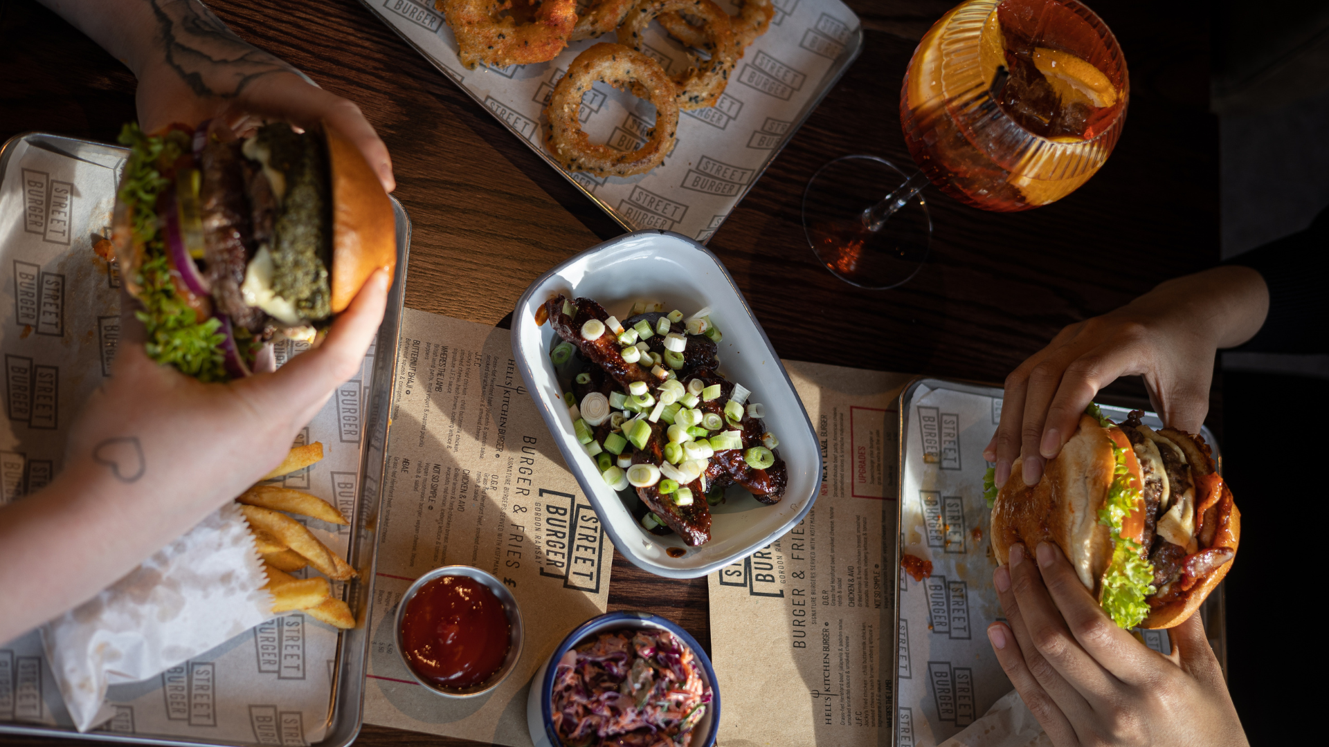 Spread of food for two people, including two burgers being held, chocolate bbq wings in middle of table, and onion rings, coleslaw, seasoned fries and cocktail on the side