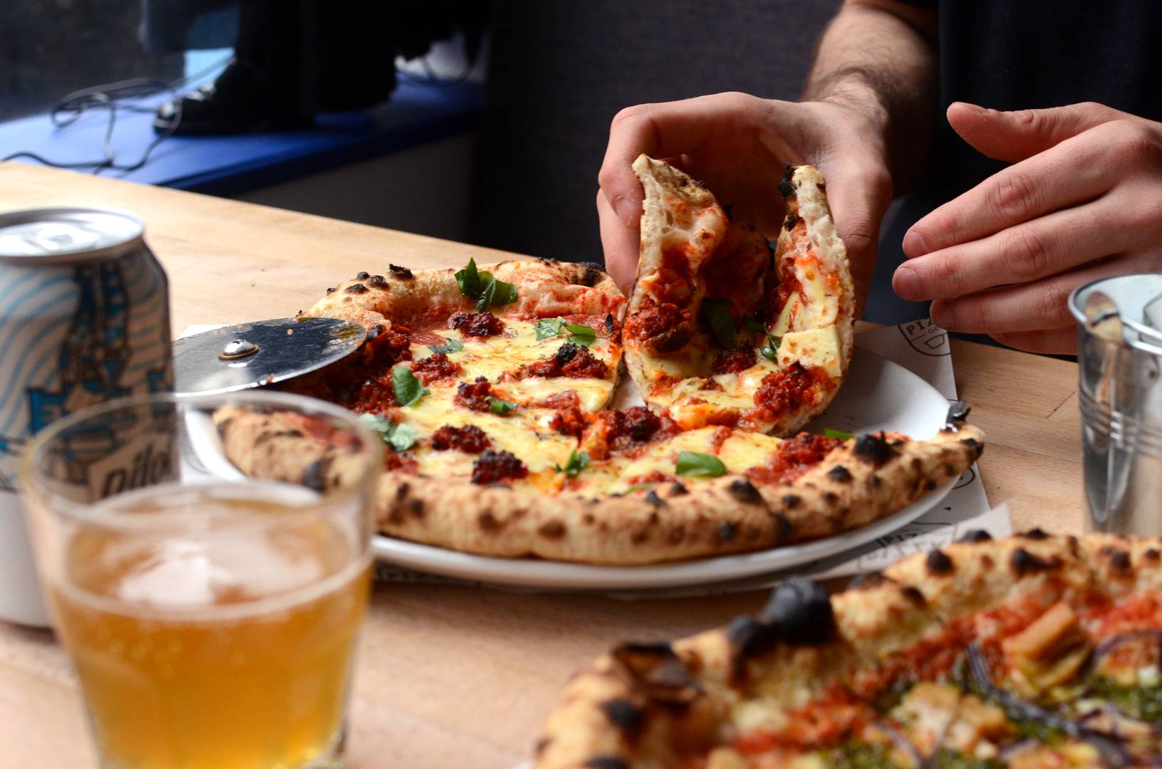A close up of a mans hand taking a slice of pizza from a plate, there is also a can and glass of beer on the table., Pizza Geeks.