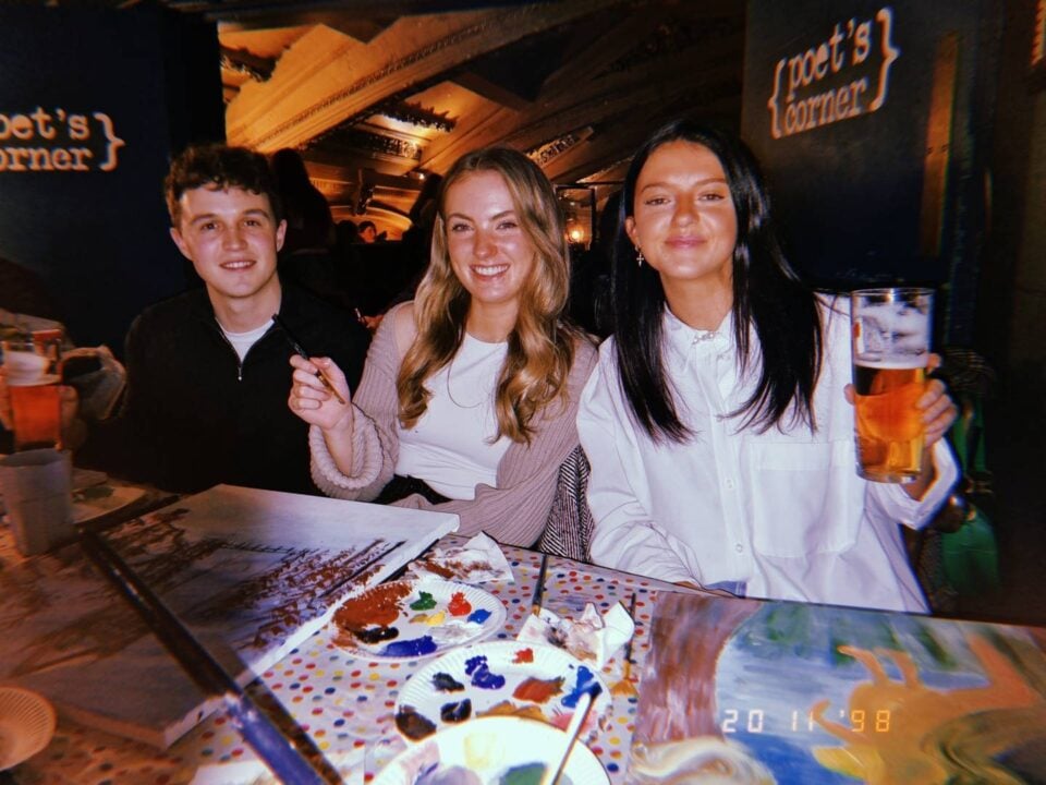 Image of 3 young people sitting in a bar with drinks but also painting pictures