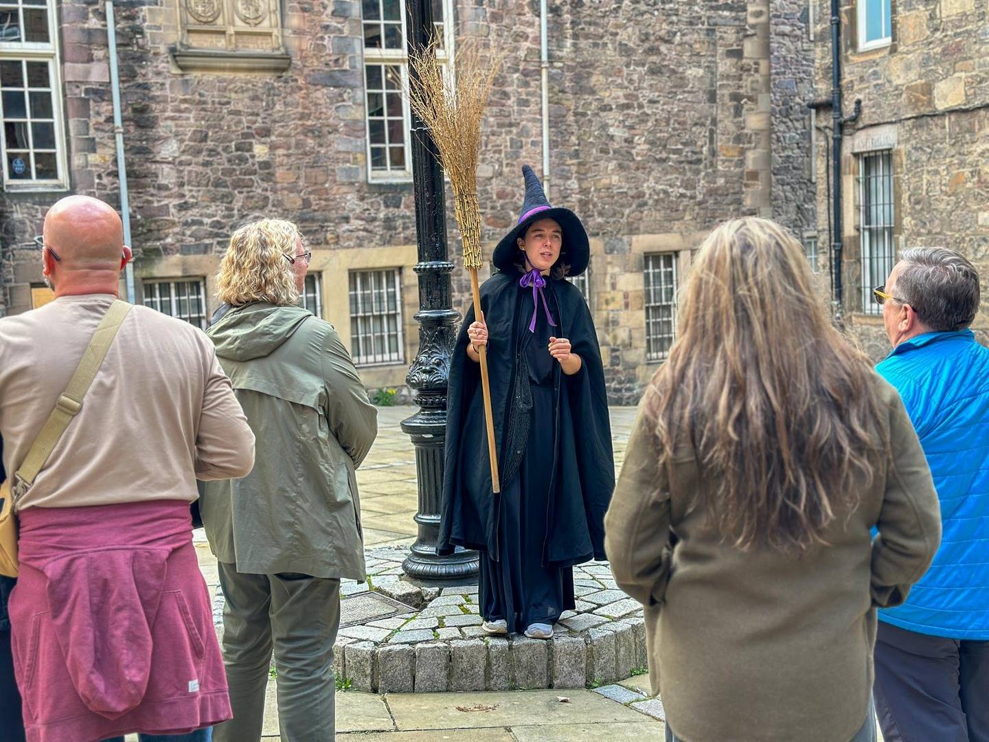 Witch guide with black pointy hat and broomstick, in a courtyard in the old town of edinburgh. Under an old style lampost.