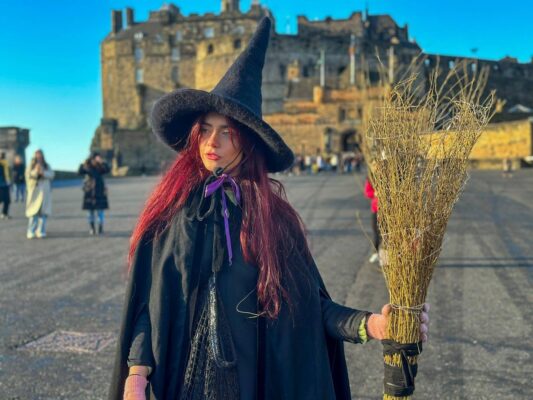 Witch guide with black pointy hat long red hair and broom stick in front of Edinburgh Castle