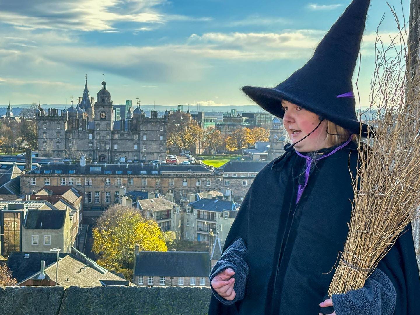 Witch guide with black pointy hat and broomstick looking at the view from Edinburgh castle, and George Herriots School which is said to be the inspiriation for Hagworts from Harry Potter.