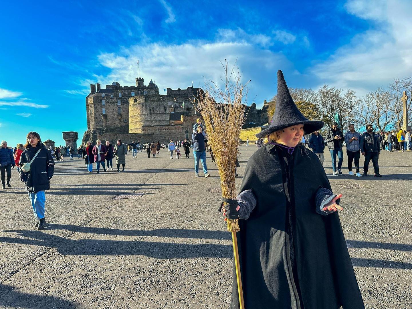 Witch guide with black pointy hat and broom stick in front of Edinburgh Castle