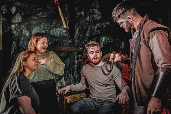 A group of visitors, one sitting in a chair, are being shown a large rusty hooked instrument by one of the performers who is dressed in medieval costume with a white and bloodied face.