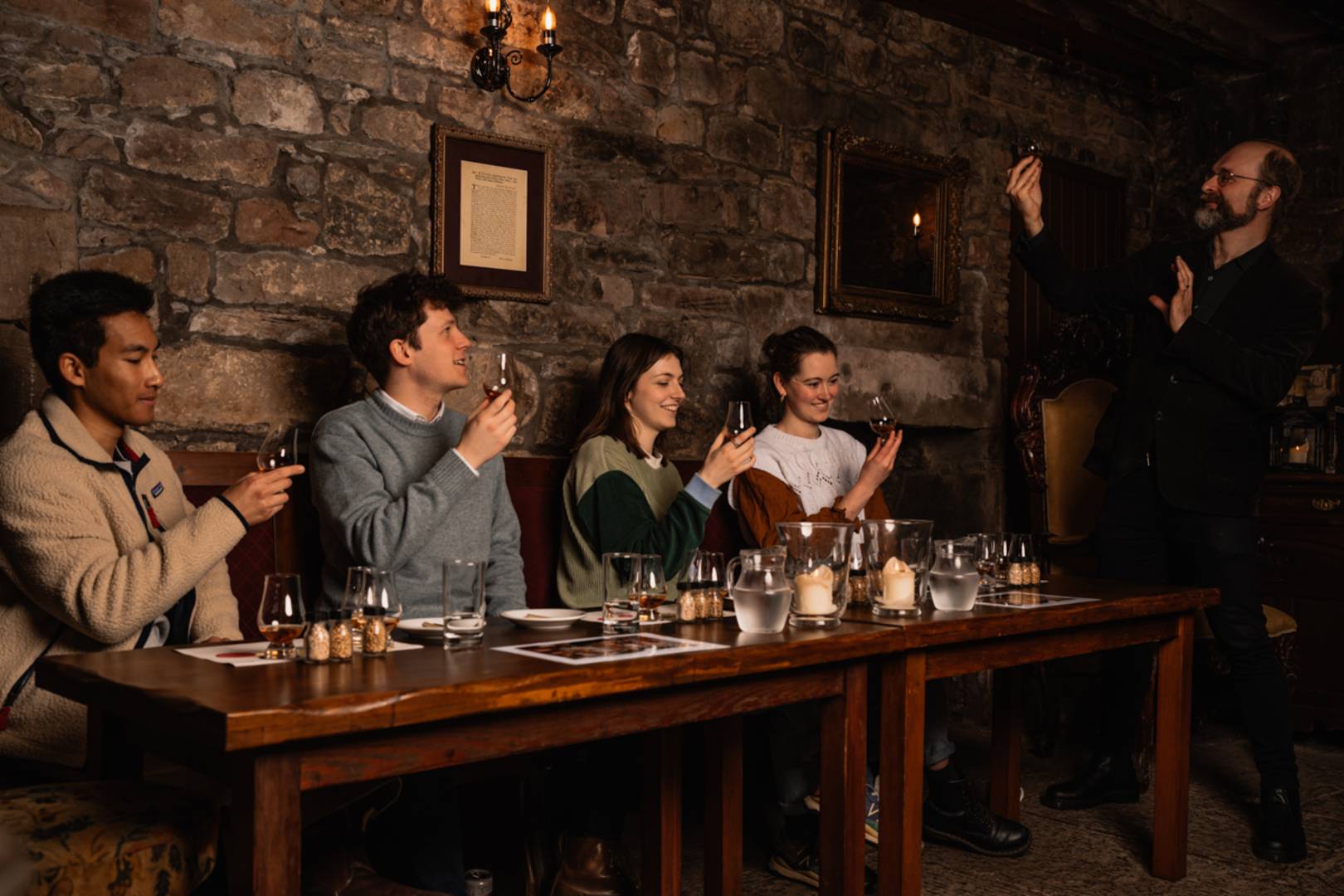 A Mercat Tours Storyteller stands next to a table of visitors in an underground cellar, and they all hold up whisky glasses.