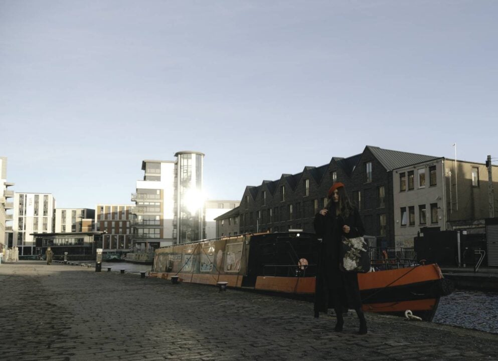 A woman is standing by the Edinburgh canal in front of a house boat with the city scape behind her. She's holding a floral printed white, gold and dark blue Georgia Victoria purse and wearing a bright red beret. ,© Georgia Victoria