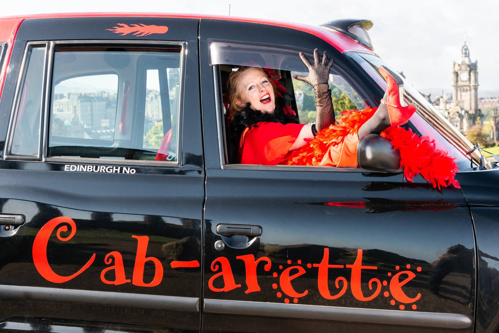 Lola in full swing. Dressed in red feathered and with a leg out the drivers window of the cabaret cab, atop Calton hill,© Kristof Turi