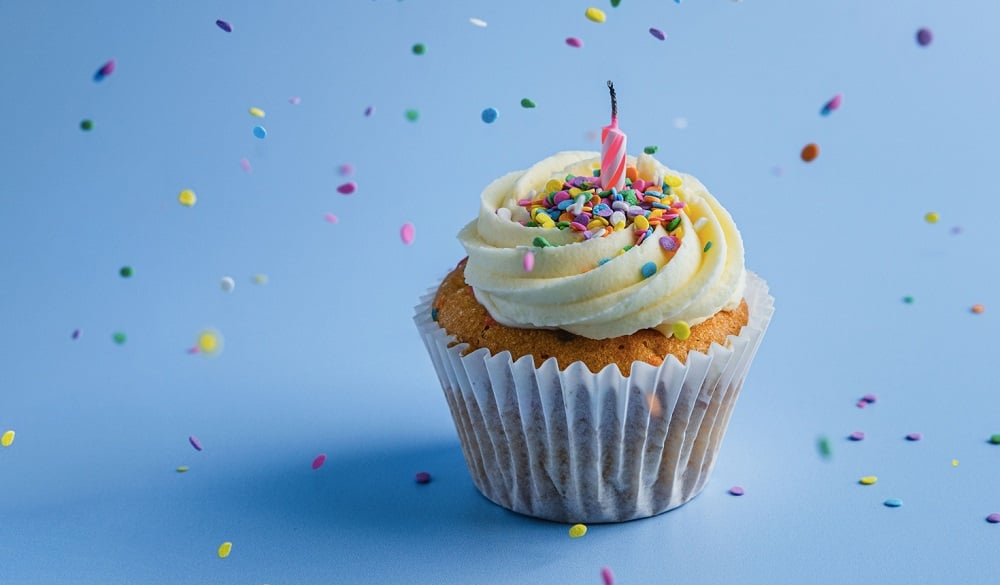A vanilla birthday cupcake with a candle in it. Multicoloured sprinkles are being sprinkled on top.