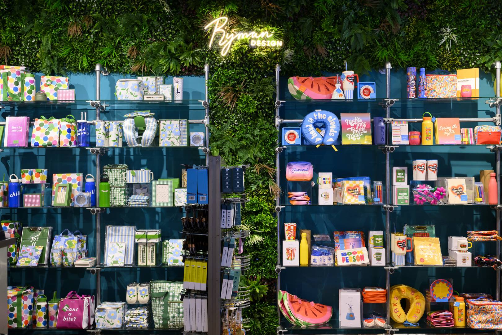 An image of a wall in Ryman Design. The wall is covered in green foliage with a lit up Ryman Design logo at the top. There is product units on the left and right displaying products such as neck pillows, notebooks and water bottles.,© All Rights Reserved