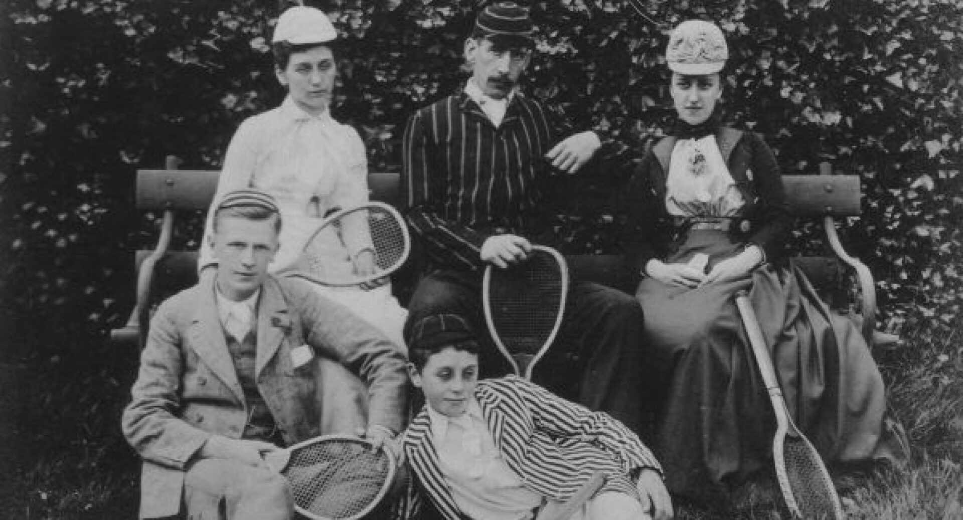 A group of five Victorian tennis players, holding racquets, sitting on a bench, and dressed in normal clothing