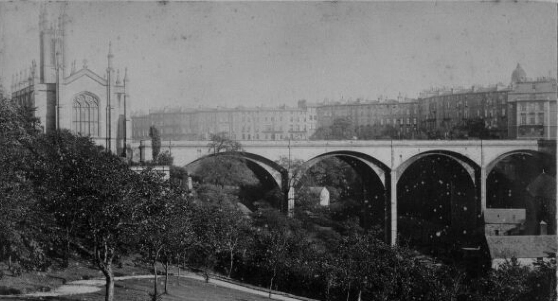 Holy Trinity Kirk and Dean Bridge, black and white photograph, 1880. Photograph taken from the pleasure gardens of Belgrave Crescent. In the background is Great Stuart Street and Randolph Crescent. Through the middle arch can be seen Greenlands Mill, demolished in the early 1900s. On the right is Lyndsay’s flour mill, demolished in the 1930s.