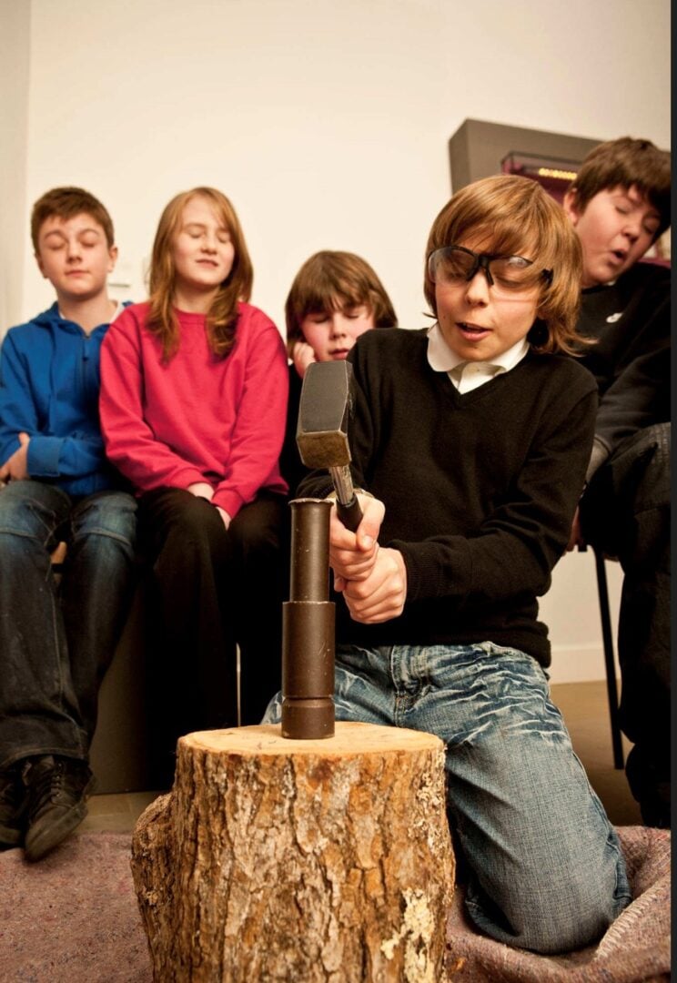 A group of children watching a young boy in safety goggles strike a coin on a log with metal implements