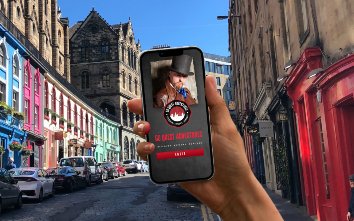 A mobile Phone being help with Go Quest Adventures on the screen.  Background of image is Victoria Street.