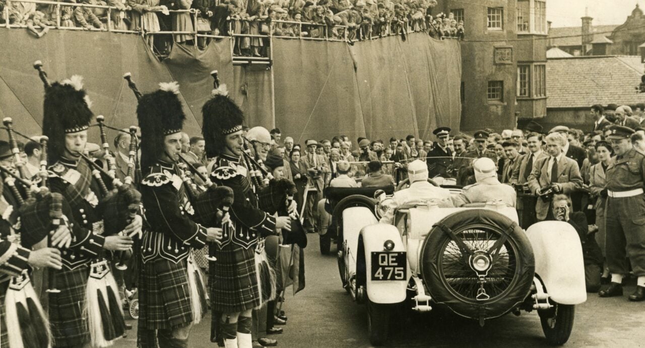 A crowd looking down on two racing cars, each with driver and passenger. Bagpipers stand to the left, more crowds to the right