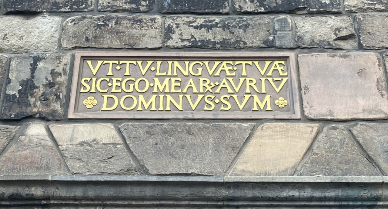 A plaque on the wall of the Museum of Edinburgh which reads in gold Latin script 'UT TU LINGUAE TUAE SIC EGO ME AR AURIU DOMINUS SUM', which means 'As thou art (master) of my tongue, so I am master of my ears'.