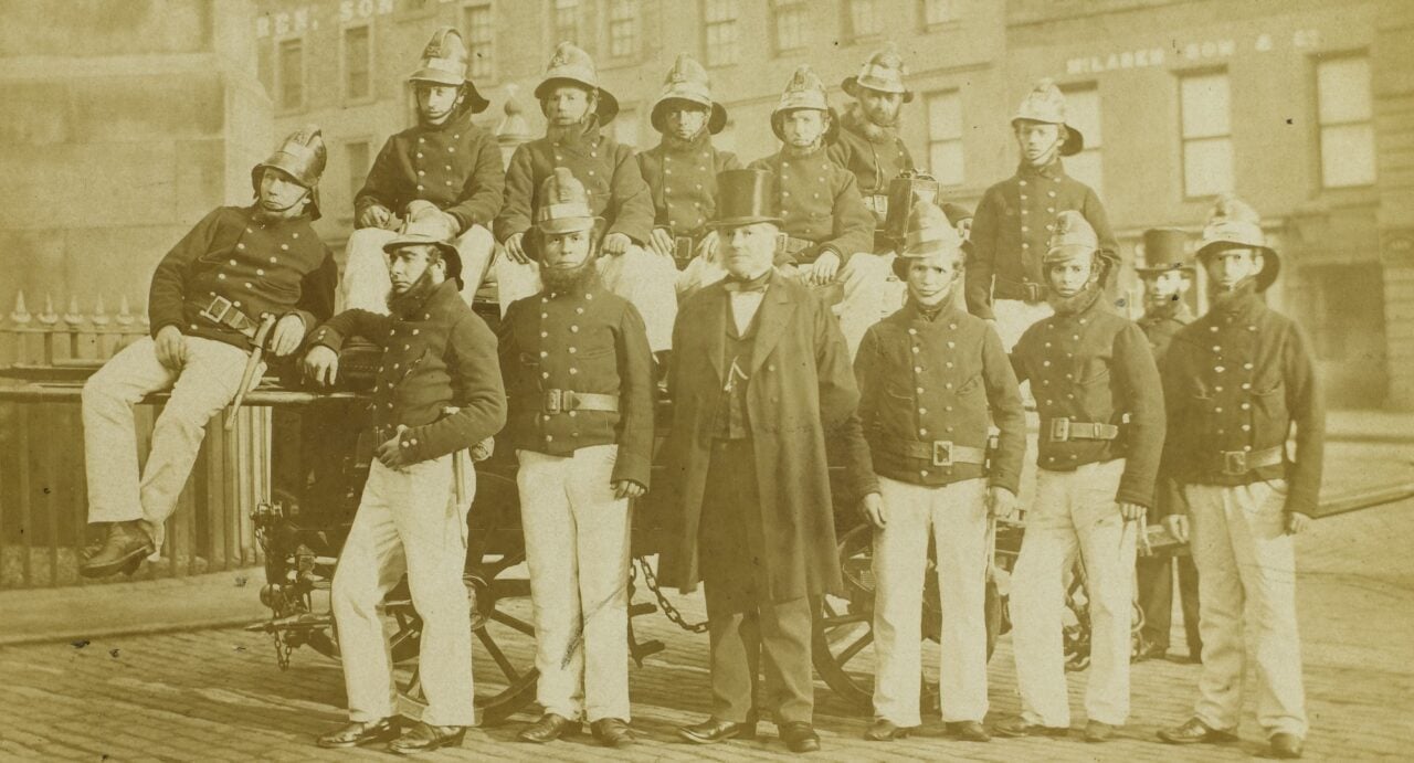 A sepia photograph of a group of firefighters in uniform standing on a cobbled street beside a man in a top hat, coat and suit