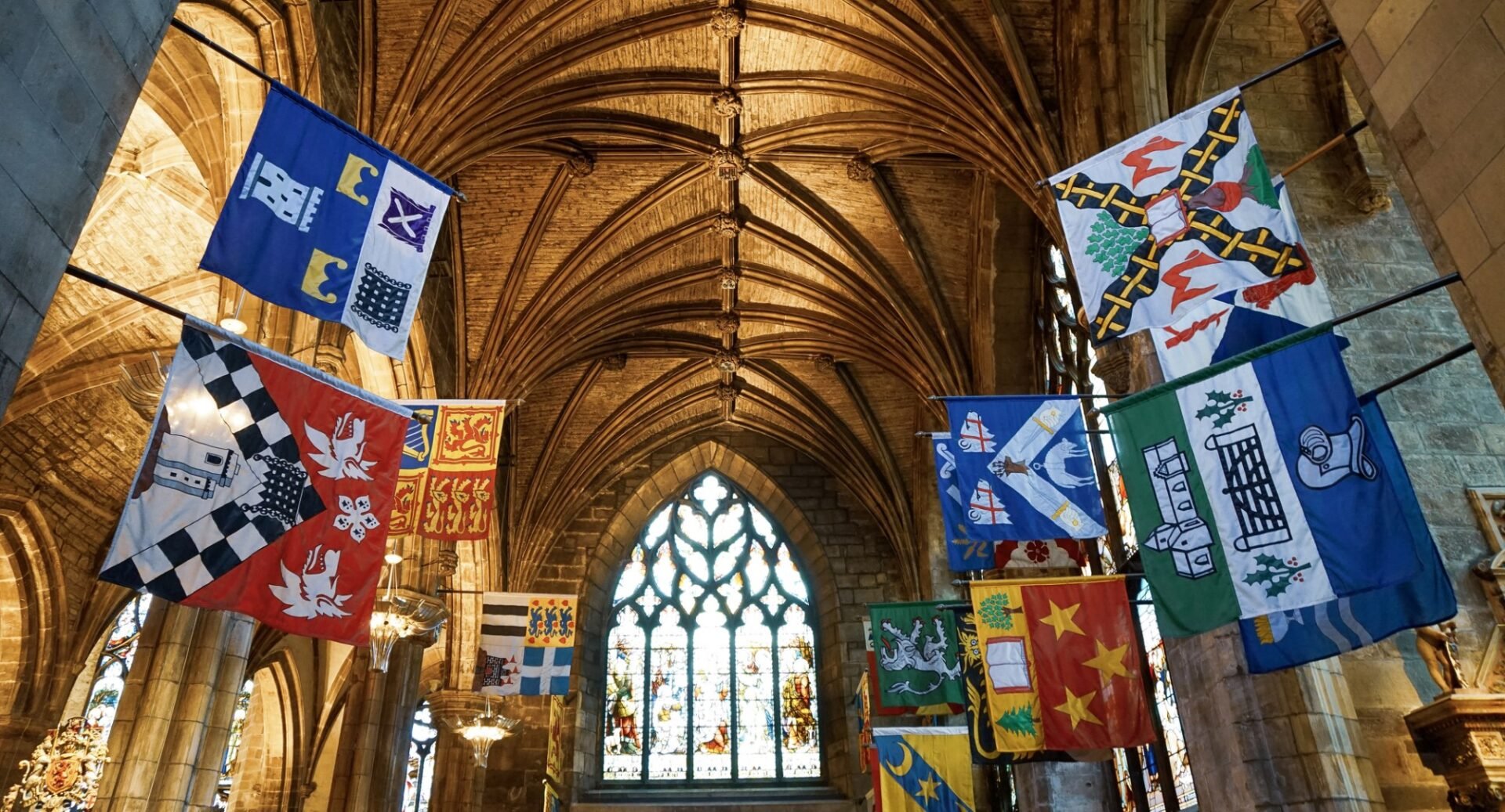 A photo of the interior of St Giles Cathedral showing a stained glass window, the vaulted ceiling, and colourful flags displayed on the pillars at the side