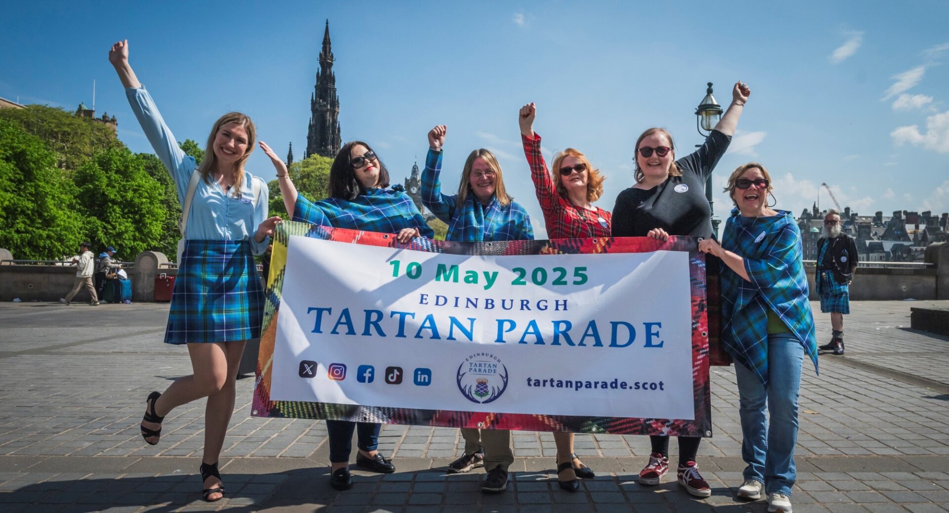 Six people with arms aloft holding a Tartan Parade banner