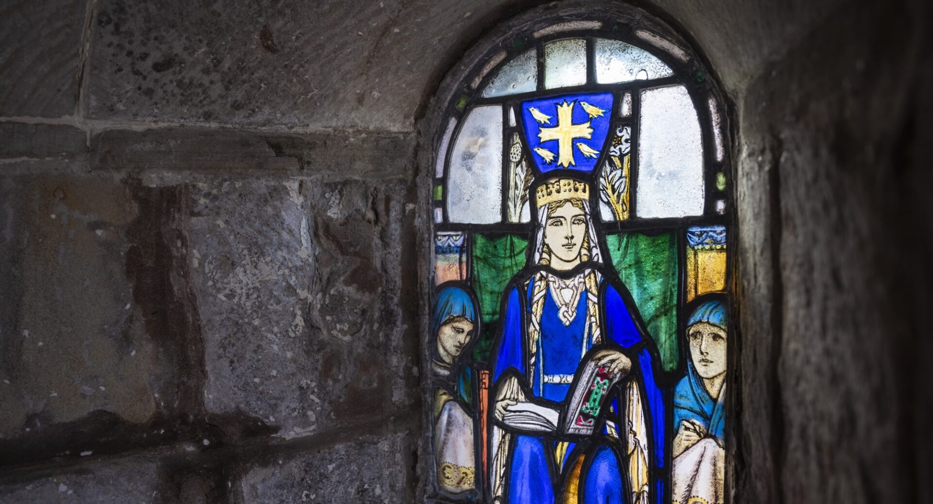 A stained glass window in St Margaret's chapel showing her dressed in blue, wearing a crown and holding a book, with two people behind her