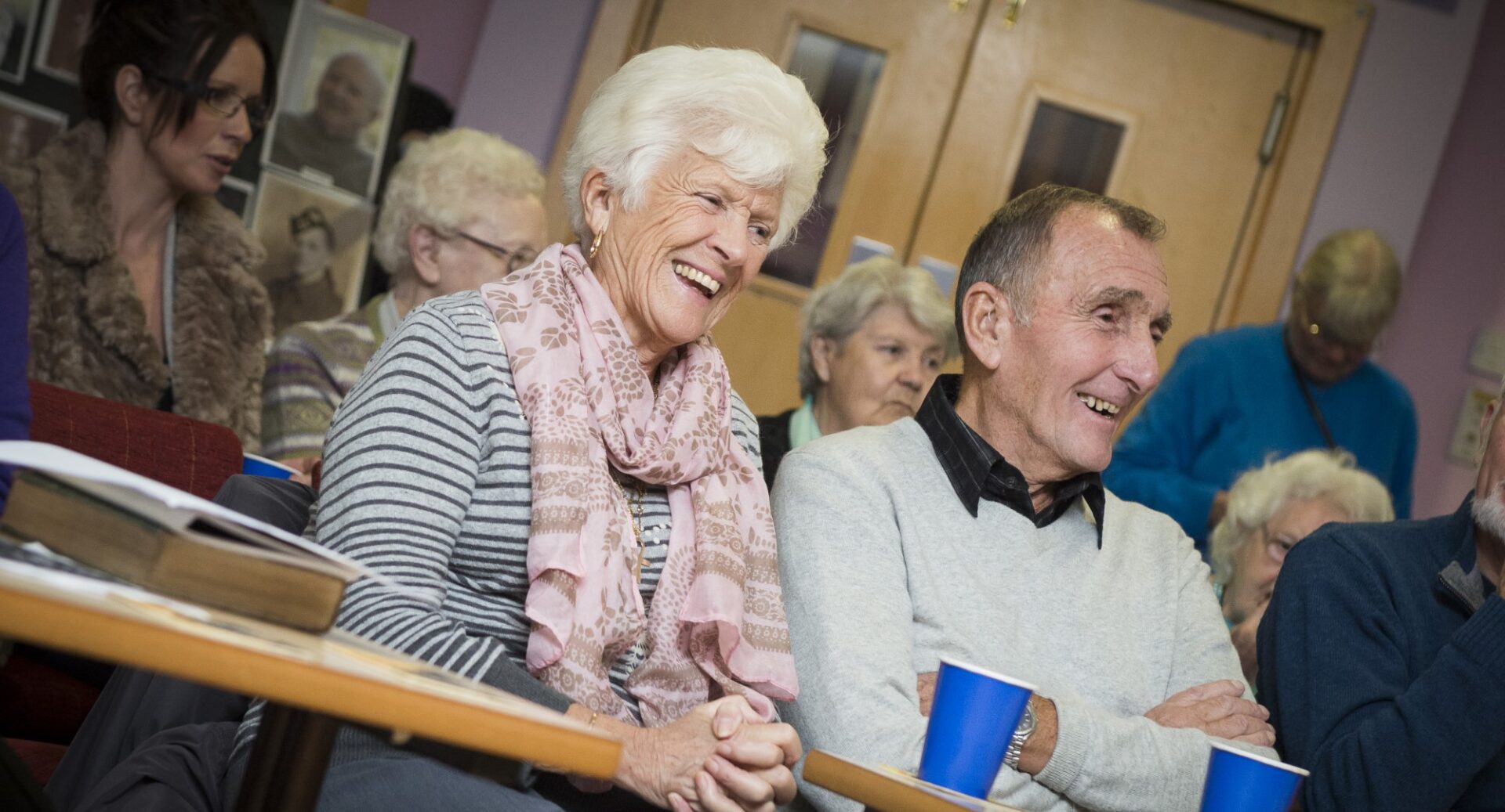 Two people sitting at a desk smiling at something off camera, other people are sitting at desks around them at a Citadel event at Leith Dockers club