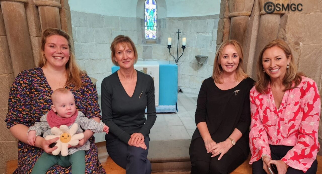 Three women, one with a baby on her lap, sitting smiling at the entrance to the chapel