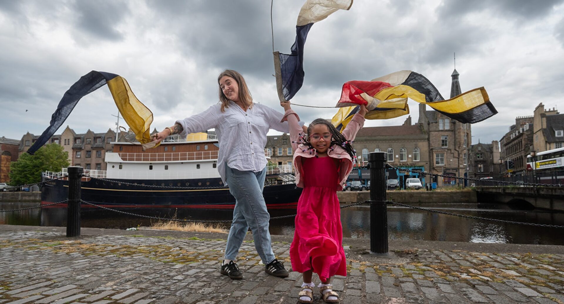 A lady in a blouse and jeans and a little girl in a red dress waving bunting on the towpath in front of the boat at Leith shore