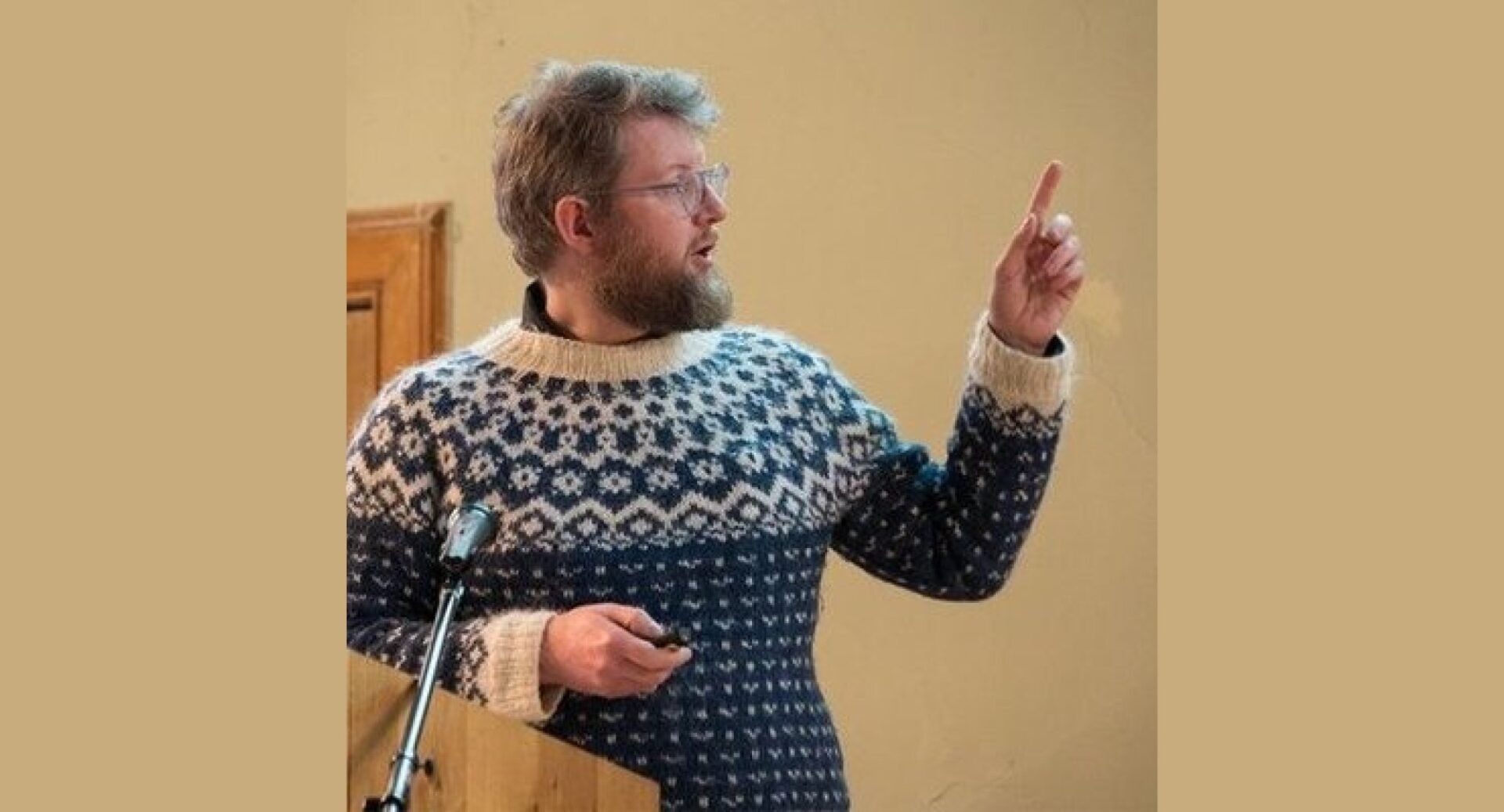 Andy Arthur standing at a lectern, wearing a Fair Isle-type jumper and pointing to a screen behind him