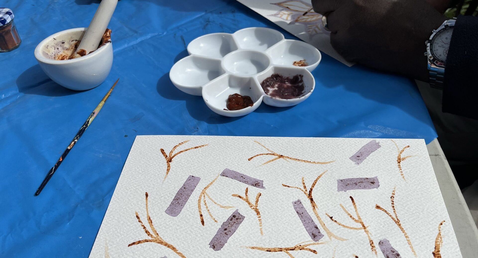 A sketch pad, a palette for paints and brushes lying on a table with a blue tablecloth