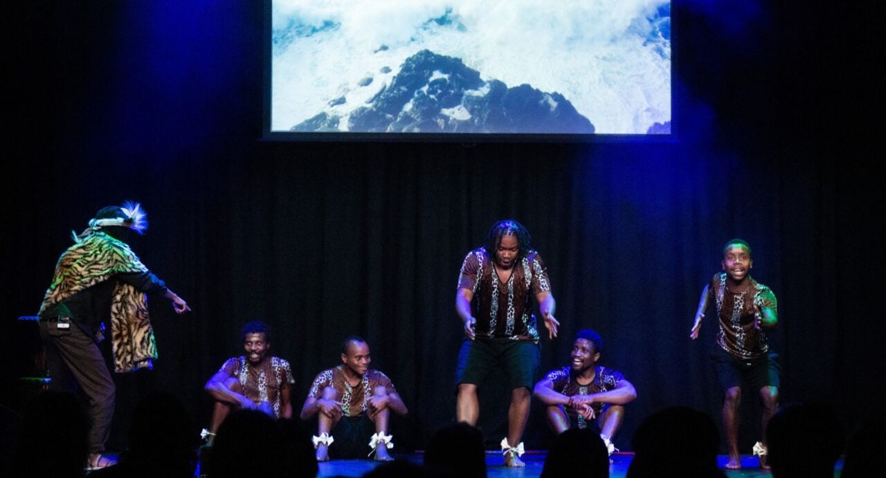 Against a backdrop of a snowy mountain five performers are in a line in the front of the stage with a sixth on the left, dressed in leopardskin, pointing at them