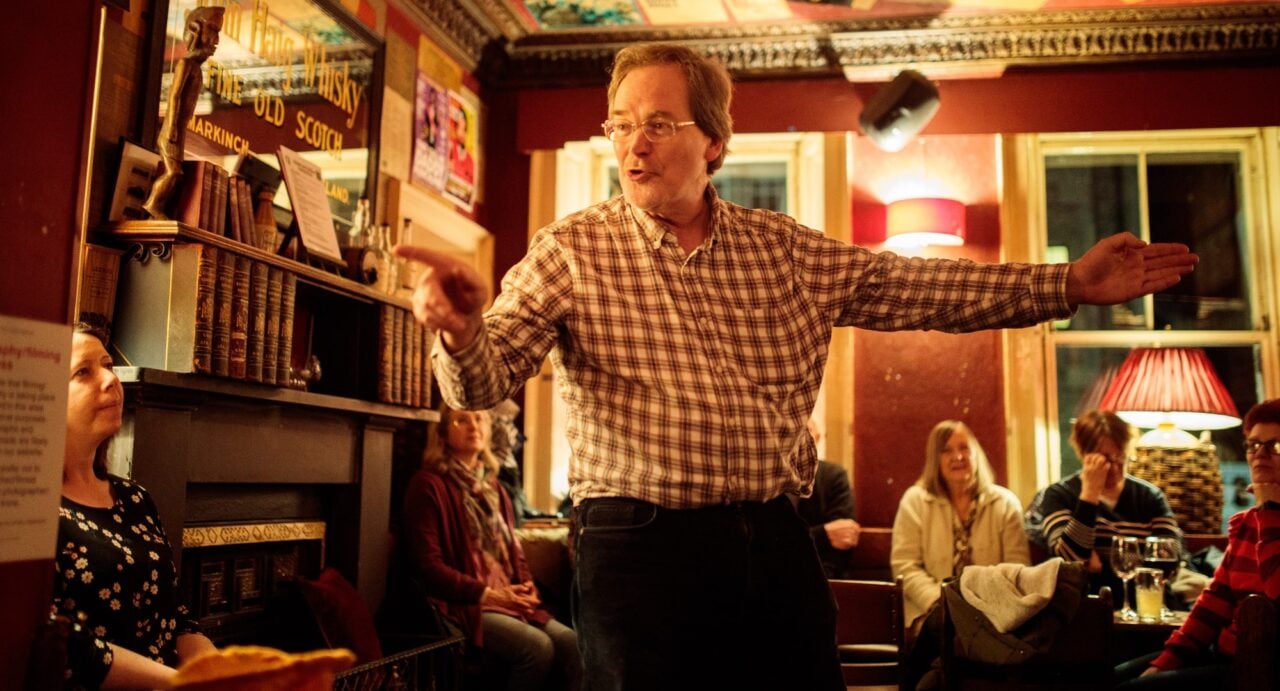 Donald Smith in a checked shirt and trousers talking animatedly to a group of women in a bar