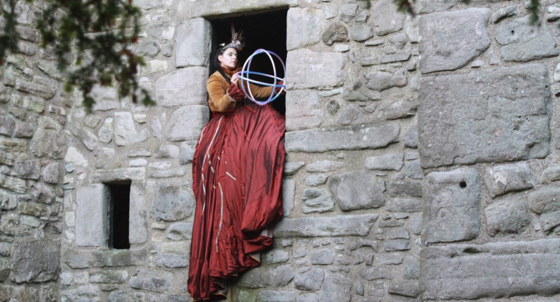 A woman in an orange mediaeval dress sitting in the stone window holding a multicoloured sphere