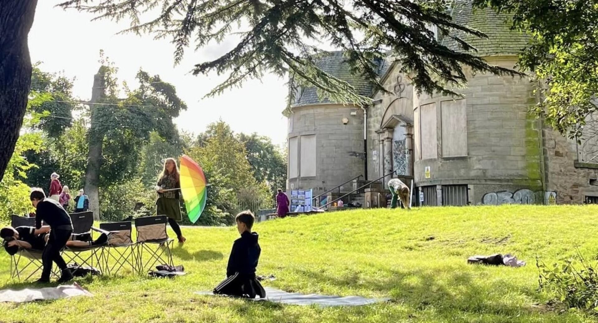 A shot under a tree of a storytelling session at Gracemount Mansion. A boy kneels on the grass, a woman holds a rainbow umbrella, other people mill round. The mansion is visible in the background.