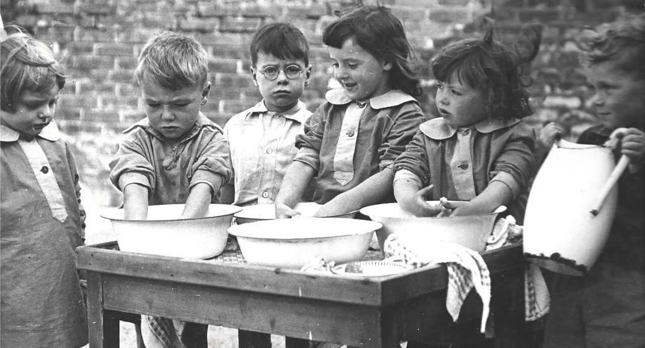 A black and white photo of primary school aged boys and girls standing by a table covered in ceramic bowls; one boy is possibly washing his hands