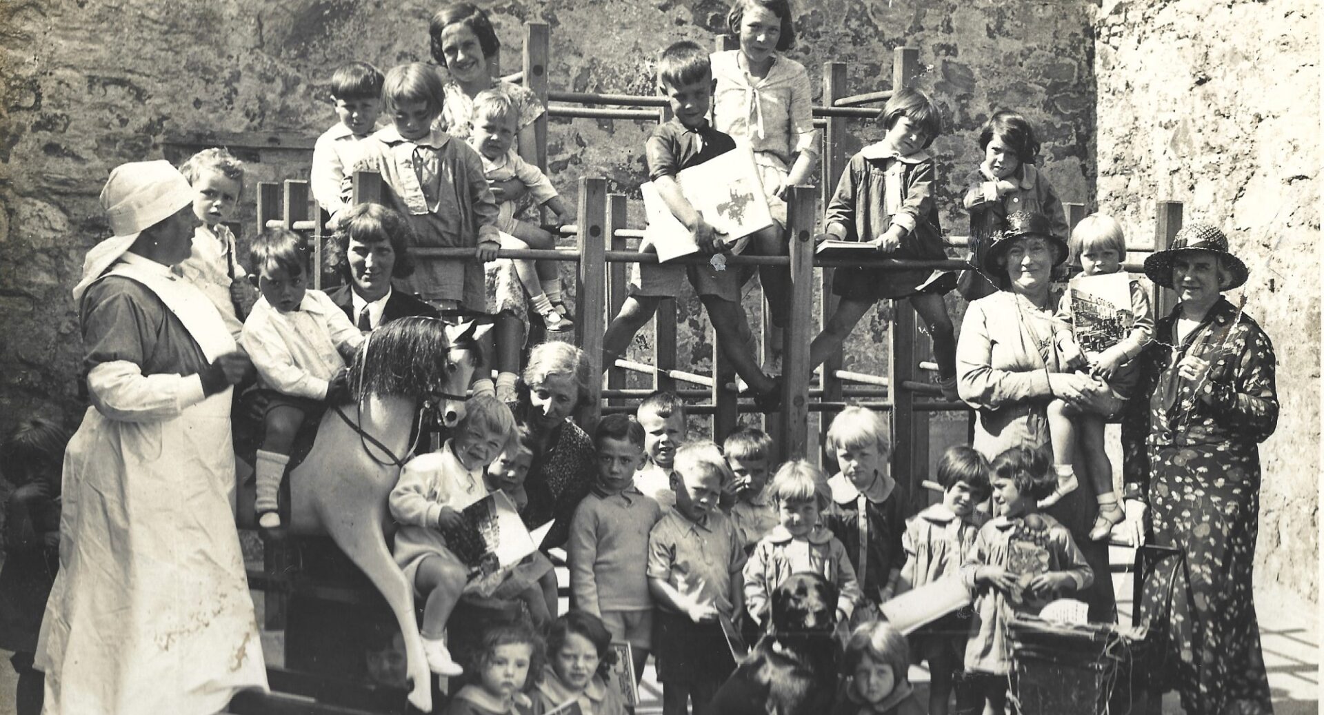 A black and white photo of a group of primary children on a climbing frame and a rocking horse with nursery staff looking on