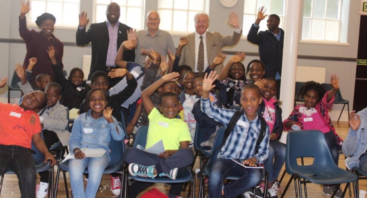 A group of waving adults and children at a Greatway event opening. The children are sitting on blue bucket seats; the adults are standing behind.