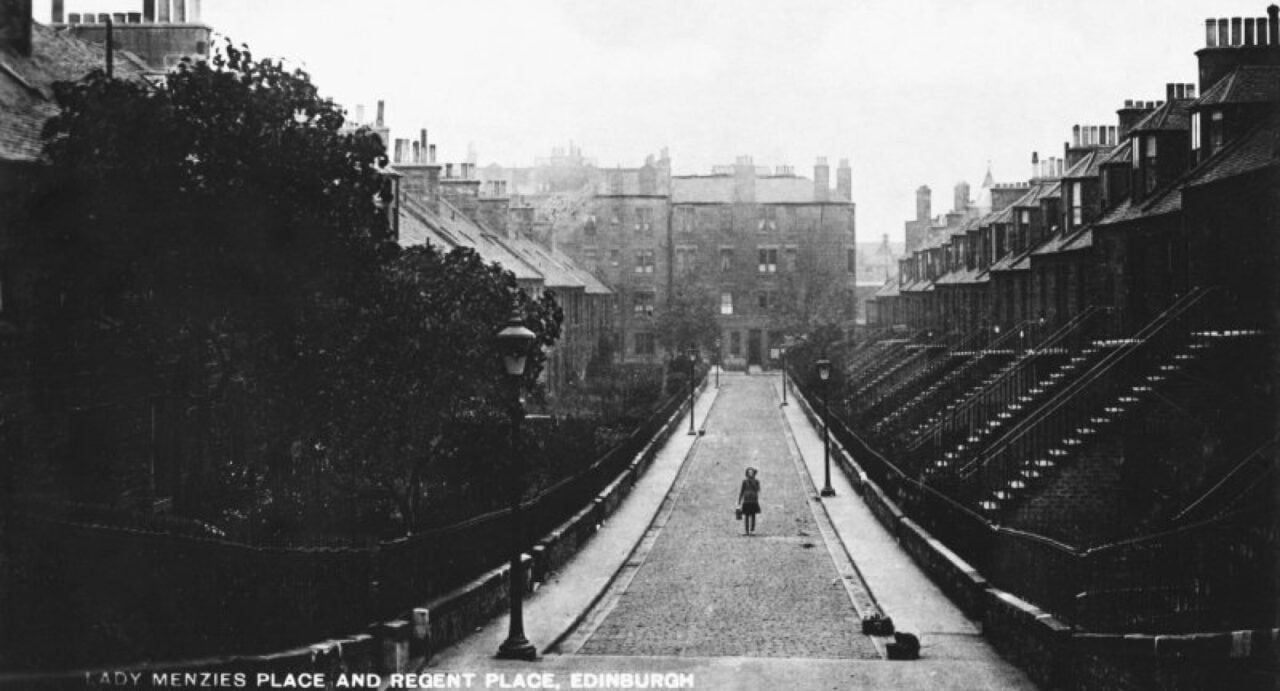 A black and white image of Lady Menzies Place and Regent Place in Edinburgh, showing a woman walking up a cobbled street with colony houses either side, and tenement flats at the far end