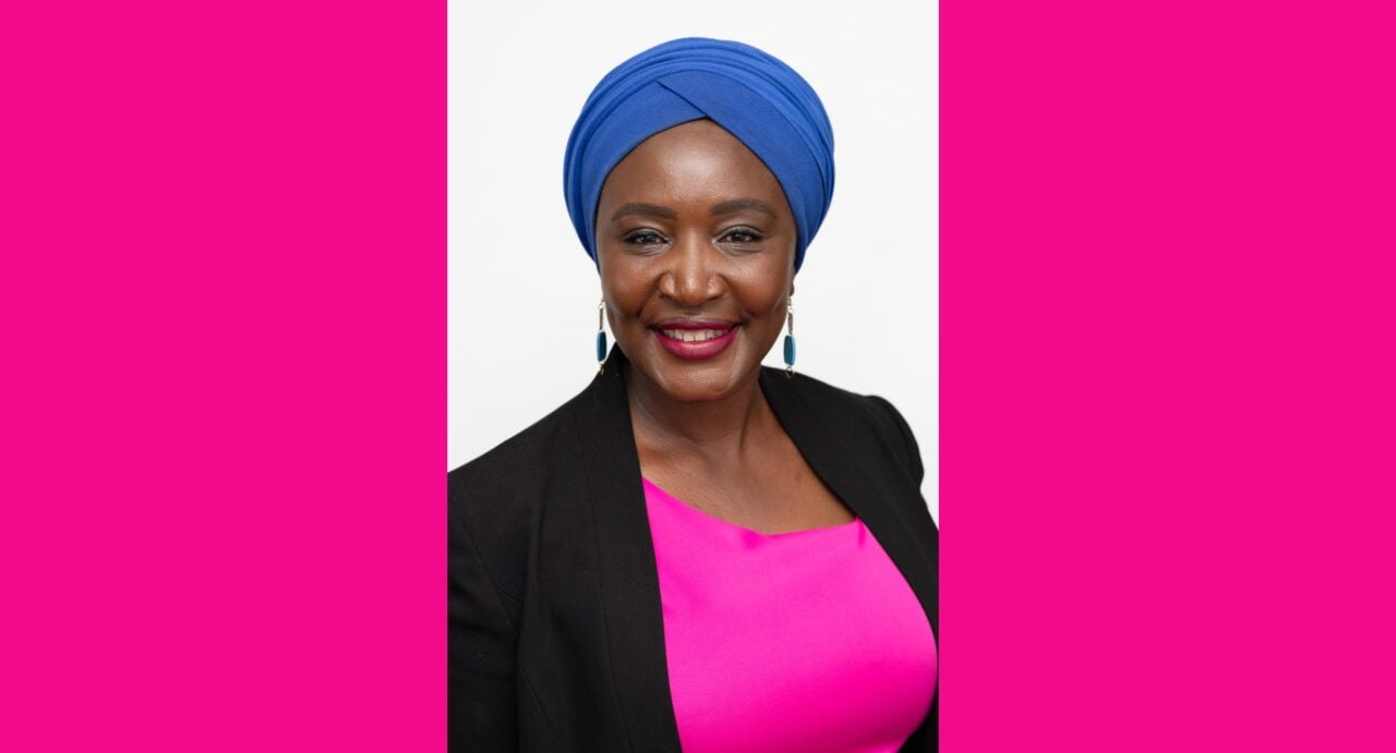 A head and shoulders portrait of Irene Mosota, wearing a pink top, black jacket, and blue head wrap