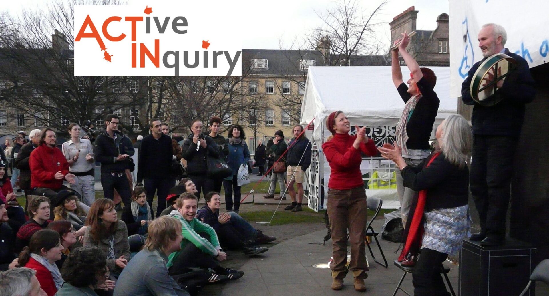 A white tent is in the background as a man stands on a box playing a drum, a woman holds up something to the crowd in front of her, and another two women watch her, clapping