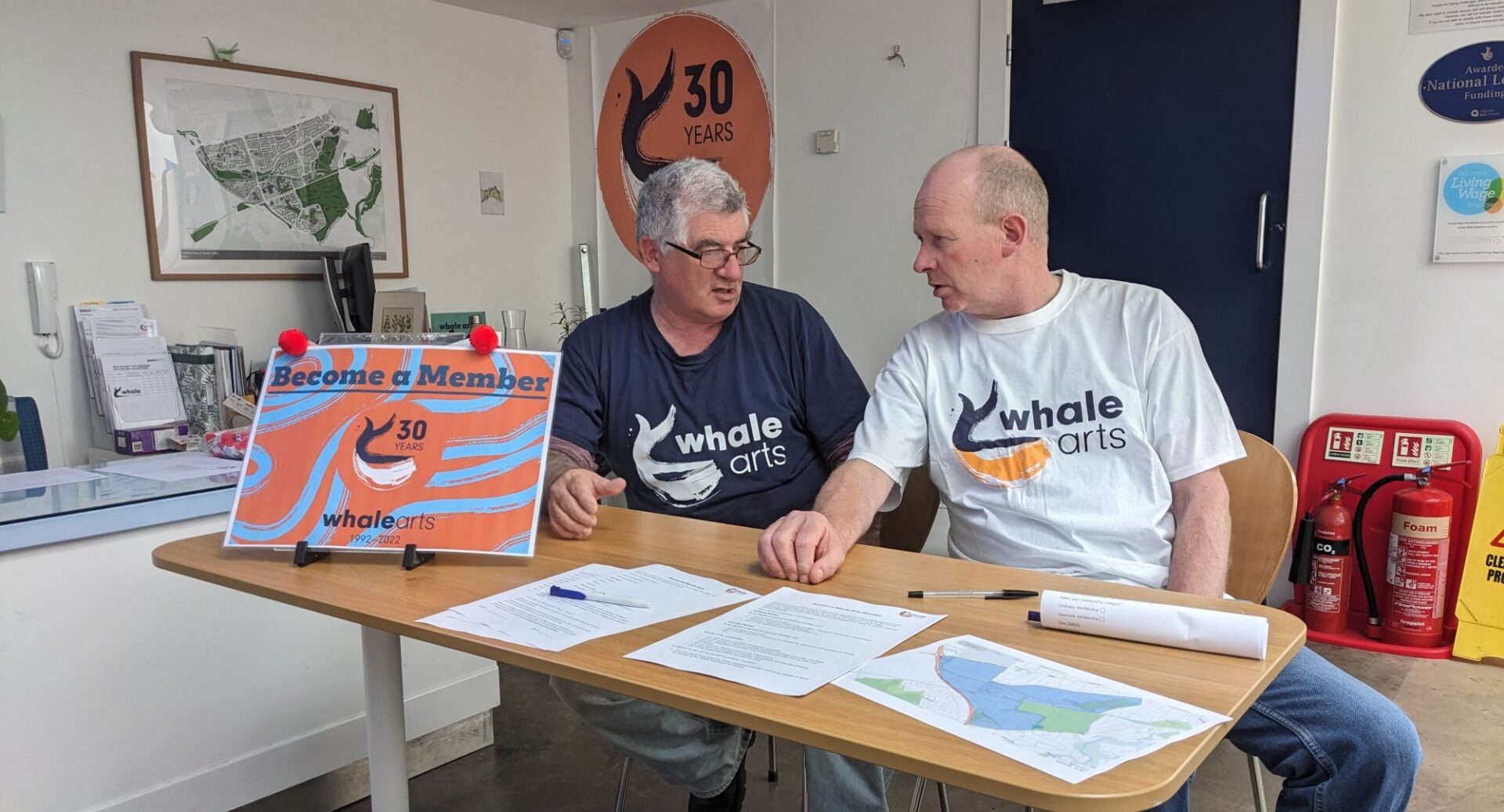 Two men, wearing WHALE Arts tshirts, sitting at a desk on which there are various sheets of paper and pens. There is artwork on the walls around them and fire extinguishers against the wall behind them