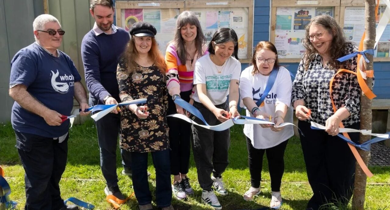 A group of people standing on grass in front of noticeboards, holding blue and white ribbons and trying to cut them