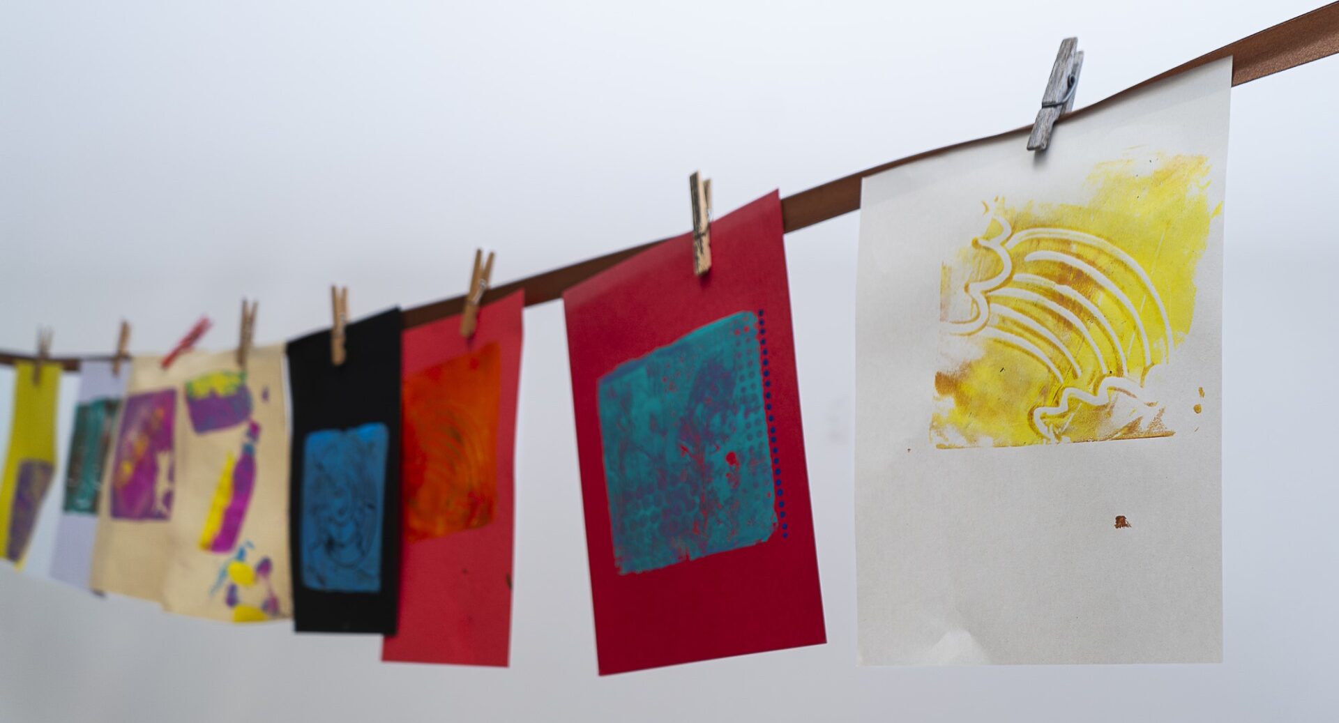 A row of abstract paintings on different coloured sheets of paper held up by clothes pegs