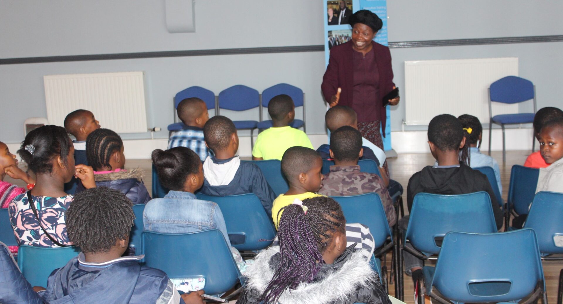 An image from the Greatway Foundation Project, Catch them young. Blue bucket chairs are laid out lecture style and several rows of young people sit listening to a woman who is standing talking at the front of the room